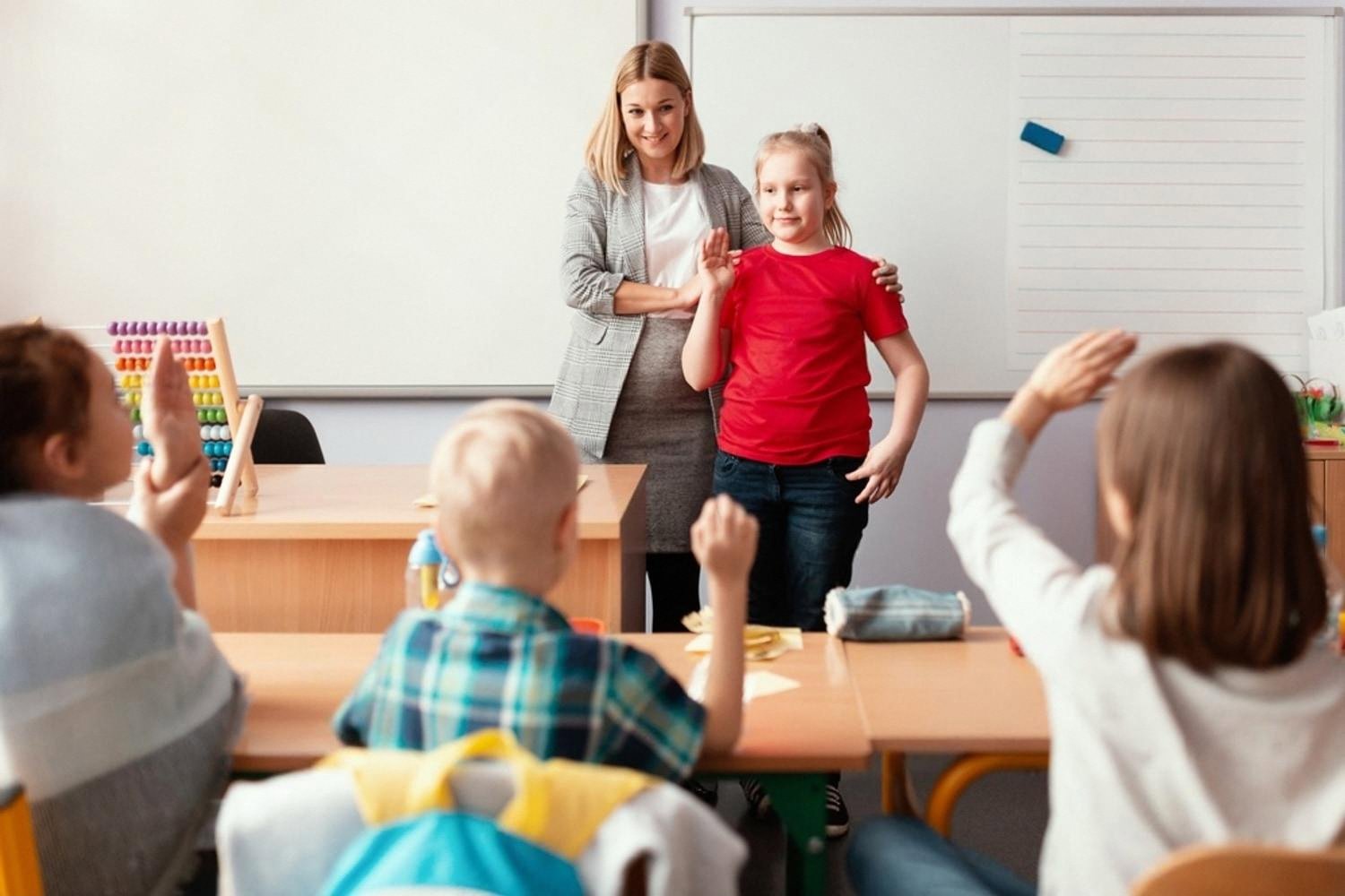 Teacher standing beside a schoolgirl at the front of a classroom while other children raise their hands